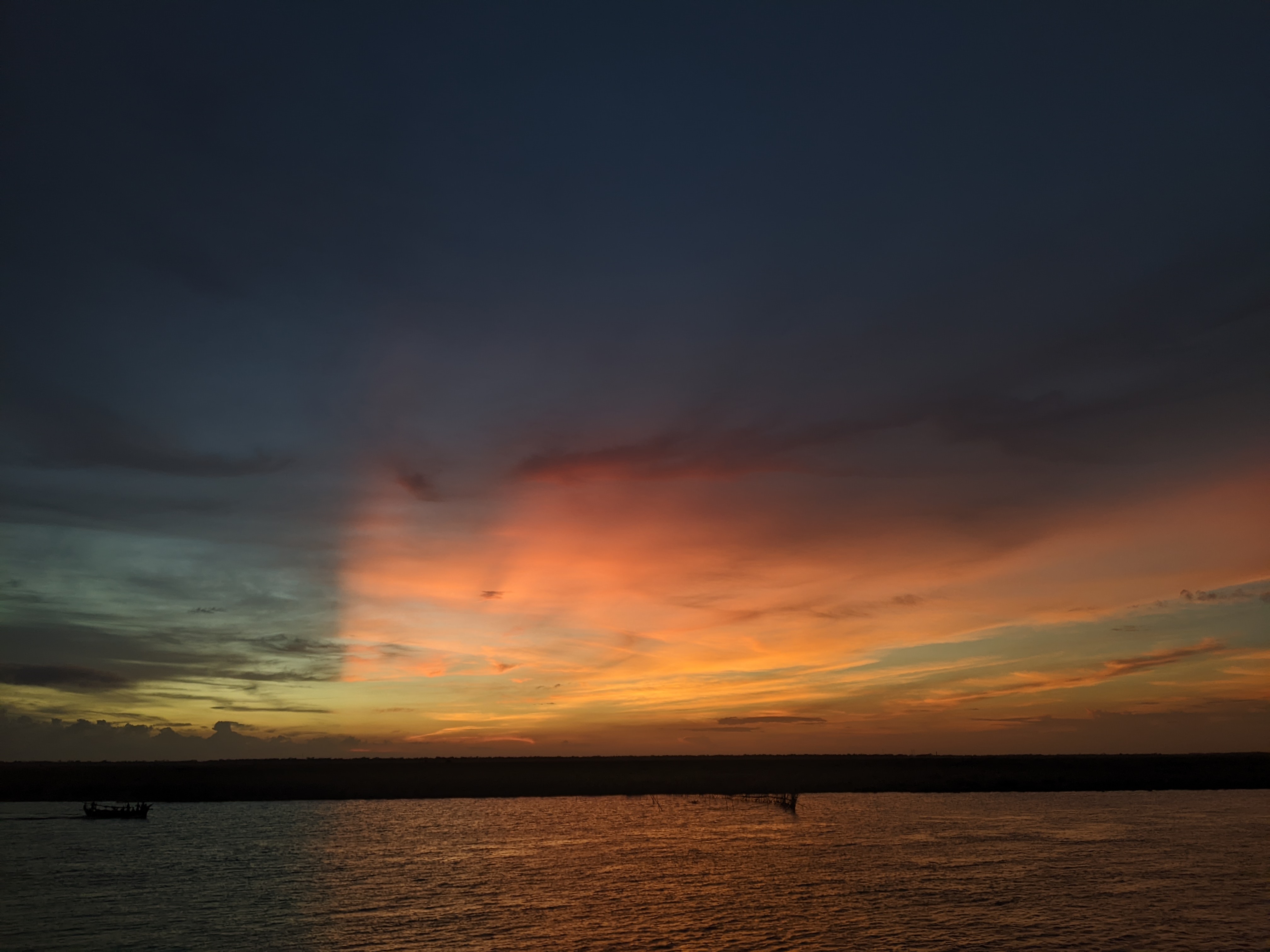 Dramatic sunset over water, dark sky above transitioning to orange and yellow hues, with a distant boat silhouette