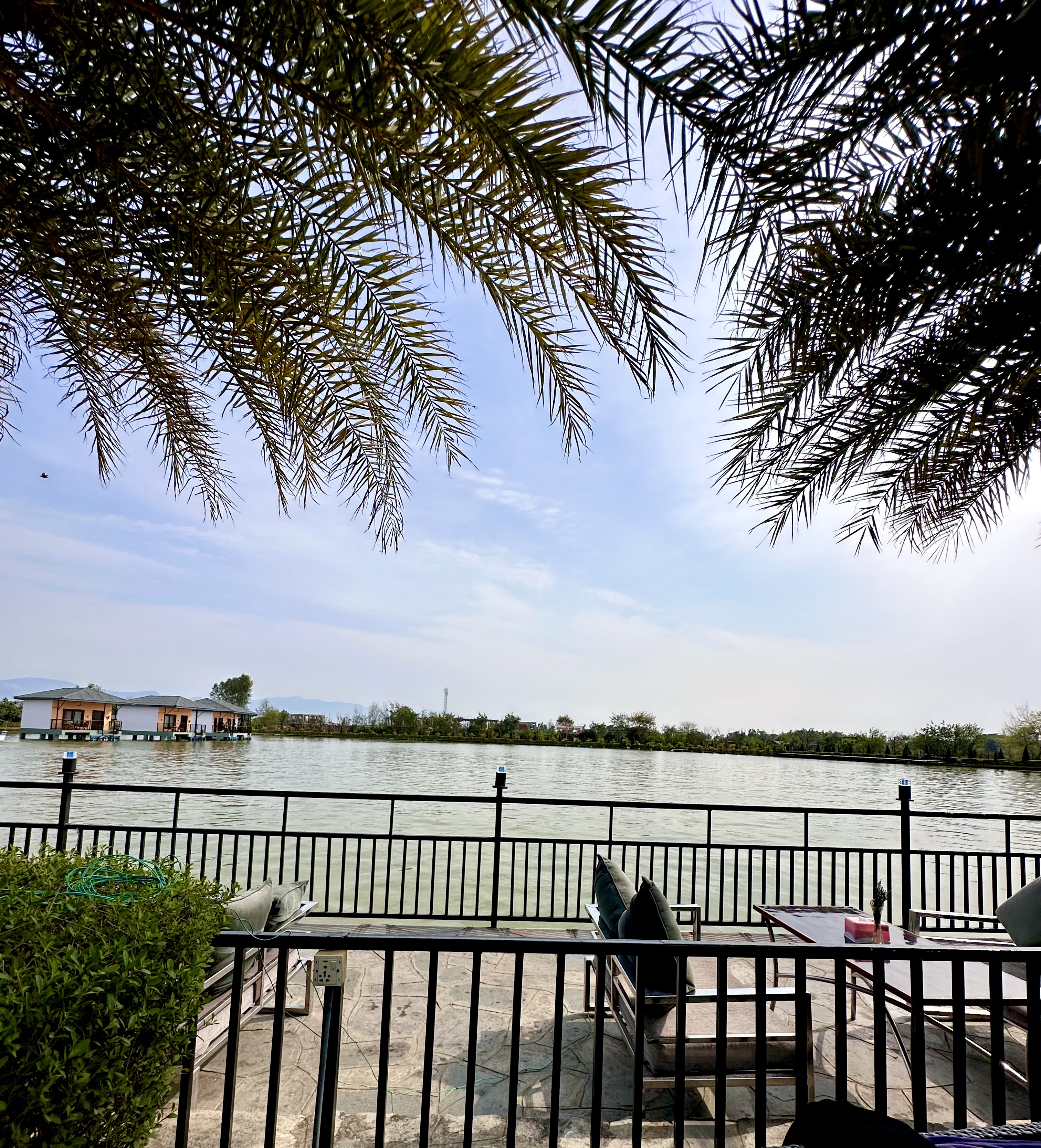 A scenic view of a lakeside area with a seating area in the foreground, consisting of a metal fence, cushioned chairs, and a table.