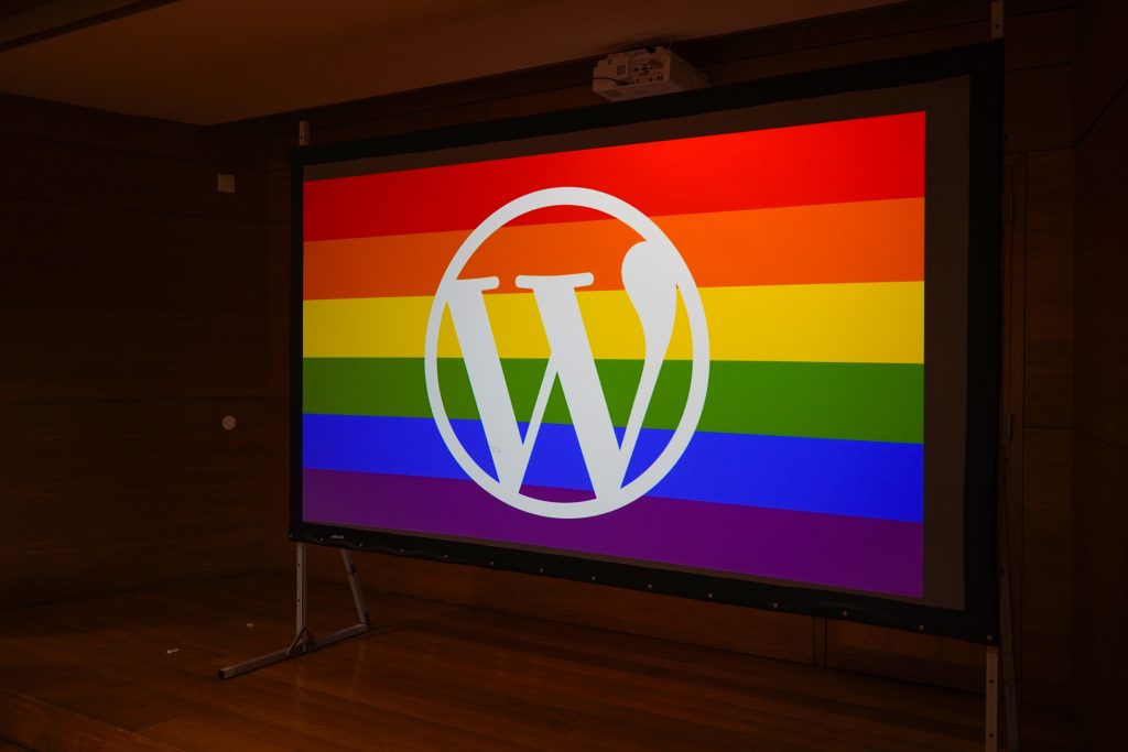 A large projection screen displays the WordPress logo in white at the center, set against a vibrant rainbow striped background. The stripes include shades of red, orange, yellow, green, blue, and purple, representing diversity and inclusivity.