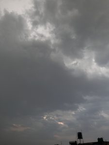 A moody sky filled with dark, dense clouds, with hints of light peeking through in some areas.The clouds range in shades of gray, suggesting an overcast atmosphere. In the foreground, silhouettes of water tanks and structures are visible against the dramatic sky.