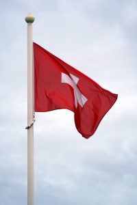 A Swiss flag is flying on a flagpole against a cloudy sky