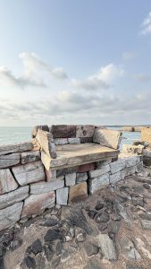 A stone structure resembling a bench is positioned on a rocky shoreline with the ocean in the background.
