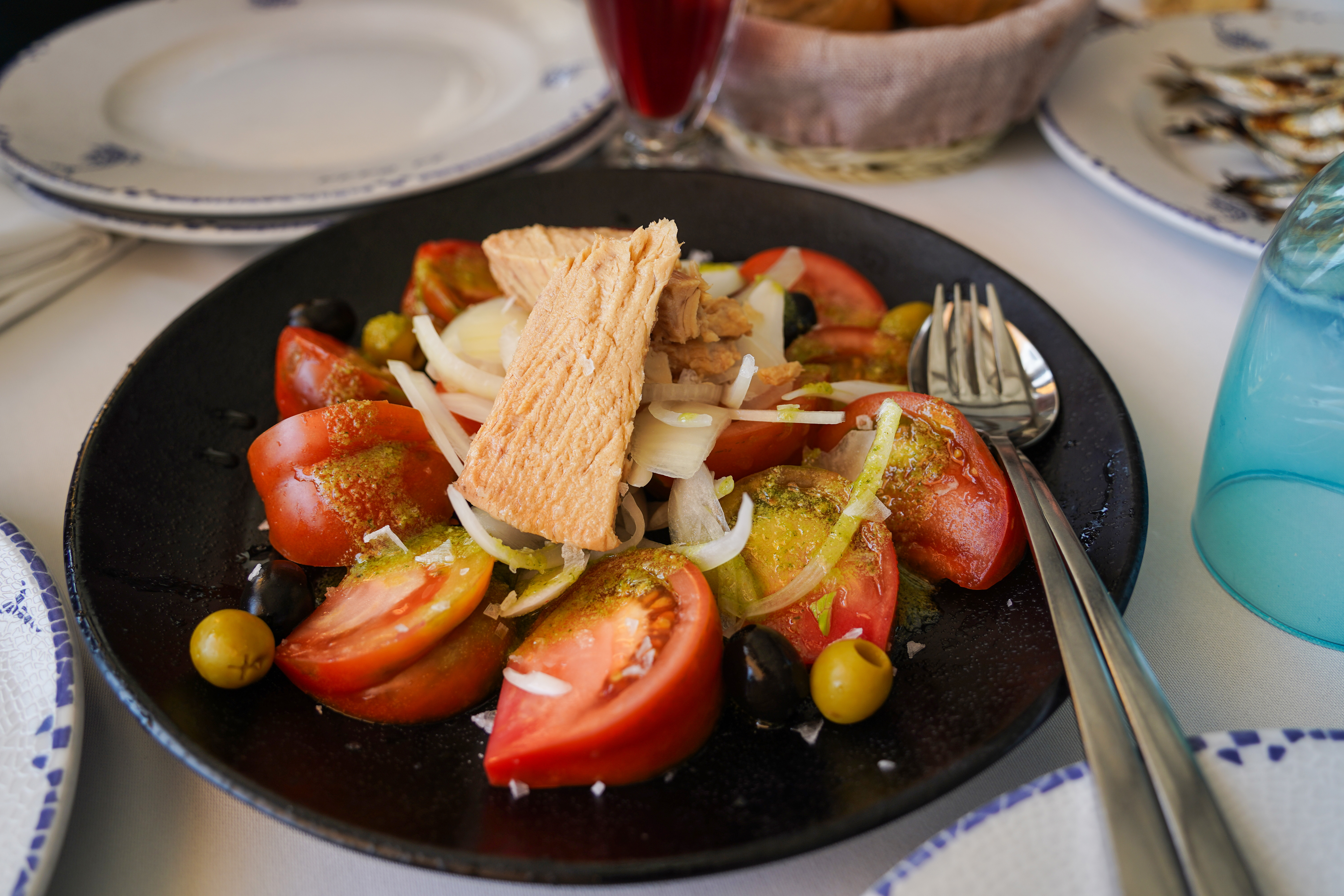 A black plate is filled with a vibrant salad featuring sliced tomatoes, olives, and onions, topped with pieces of canned tuna. The salad is garnished with a light dressing, possibly made with herbs.