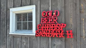 A small window set into a weathered rustic wooden wall with the words Cold Beer Cheese Sandwich painted in red, arranged vertically next to it.