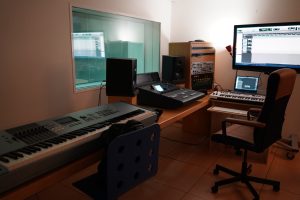 A modern music production studio featuring two keyboards on a desk, a computer monitor displaying a digital audio workstation, studio monitors on either side of the desk, and additional audio equipment in the background.