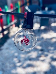 A close-up image of a decorative lightbulb hanging from a black wire, featuring a splash of red paint on its surface