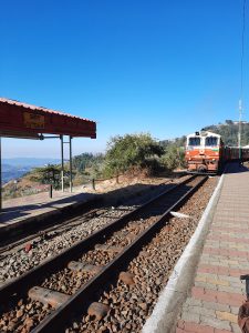 A scenic view of a small railway station under a clear blue sky. In the foreground, railway tracks lined with gravel show a train approaching the platform.
