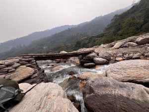 A stone bridge made of wooden logs crosses a shallow, rushing river. The river is surrounded by smooth, large stones and pebbles.