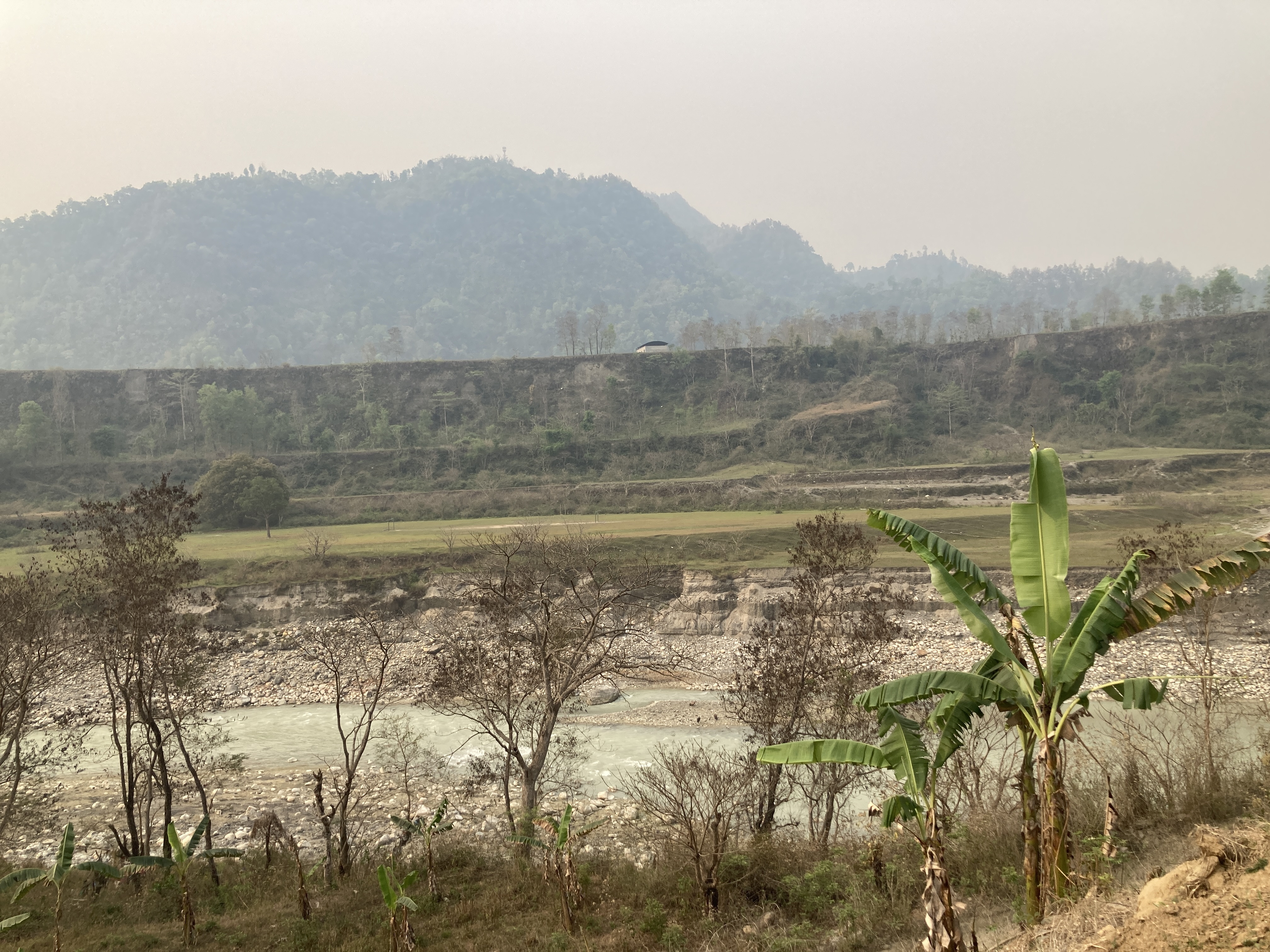 A landscape scene featuring a river winding through a rocky bed, surrounded by sparse vegetation and some banana plants in the foreground.