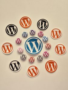 A collection of circular stickers and buttons featuring the WordPress logo in various colors, including orange, black, blue, purple, pink, and white, arranged on a light background. 
