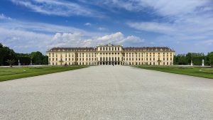Frontal view of Schönbrunn Palace in Vienna on a sunny day, with a blue sky and scattered clouds. A wide gravel path and symmetrical gardens lead to the grand yellow baroque building with numerous windows and decorative details