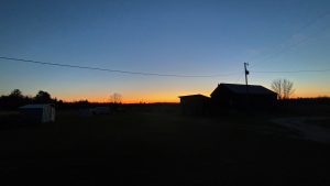 An evening sky with the glow of the setting sun on the horizon.  Silhouetted are a barn and some other farm buildings.