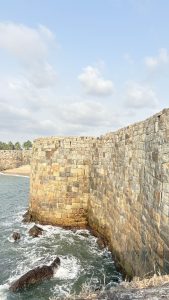 A stone fort wall rises above the water, with waves crashing against its base. The structure is made of large, uneven stones arranged in a rugged pattern.
