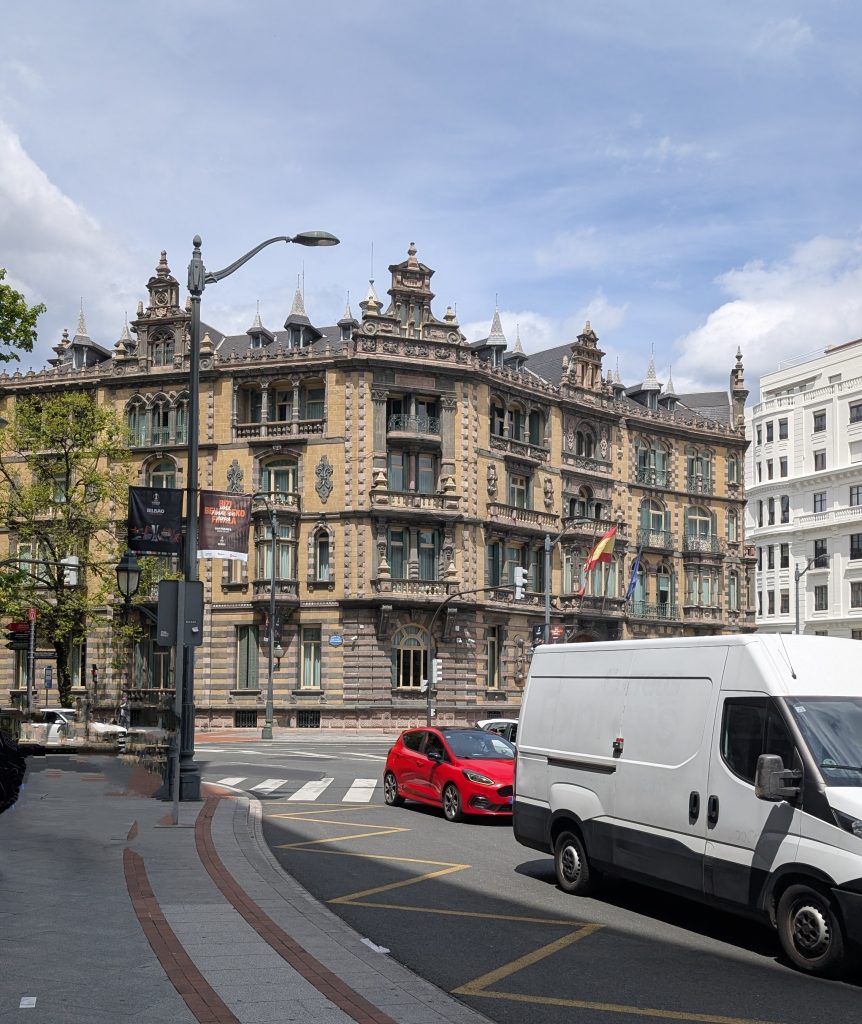 A historic, elaborately designed building stands at the corner of a street. The architecture features ornate details, multiple levels, and decorative elements.