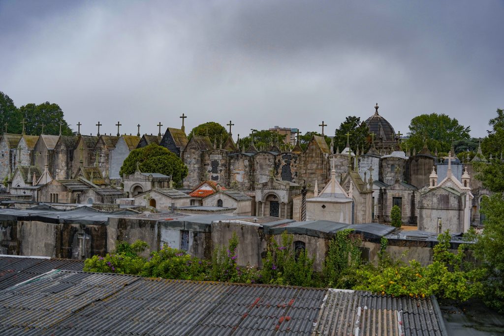 A wide view a cemetery in Porto, featuring an array of tombstones and mausoleums with elaborate designs. The structures vary in style, some with pointed roofs and ornate details, while others are more simplistic. Numerous crosses are visible atop many of the tombs, and there is a backdrop of green trees and a cloudy sky.