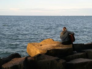 A person sitting on a large rock near the water, playing an acoustic guitar.

