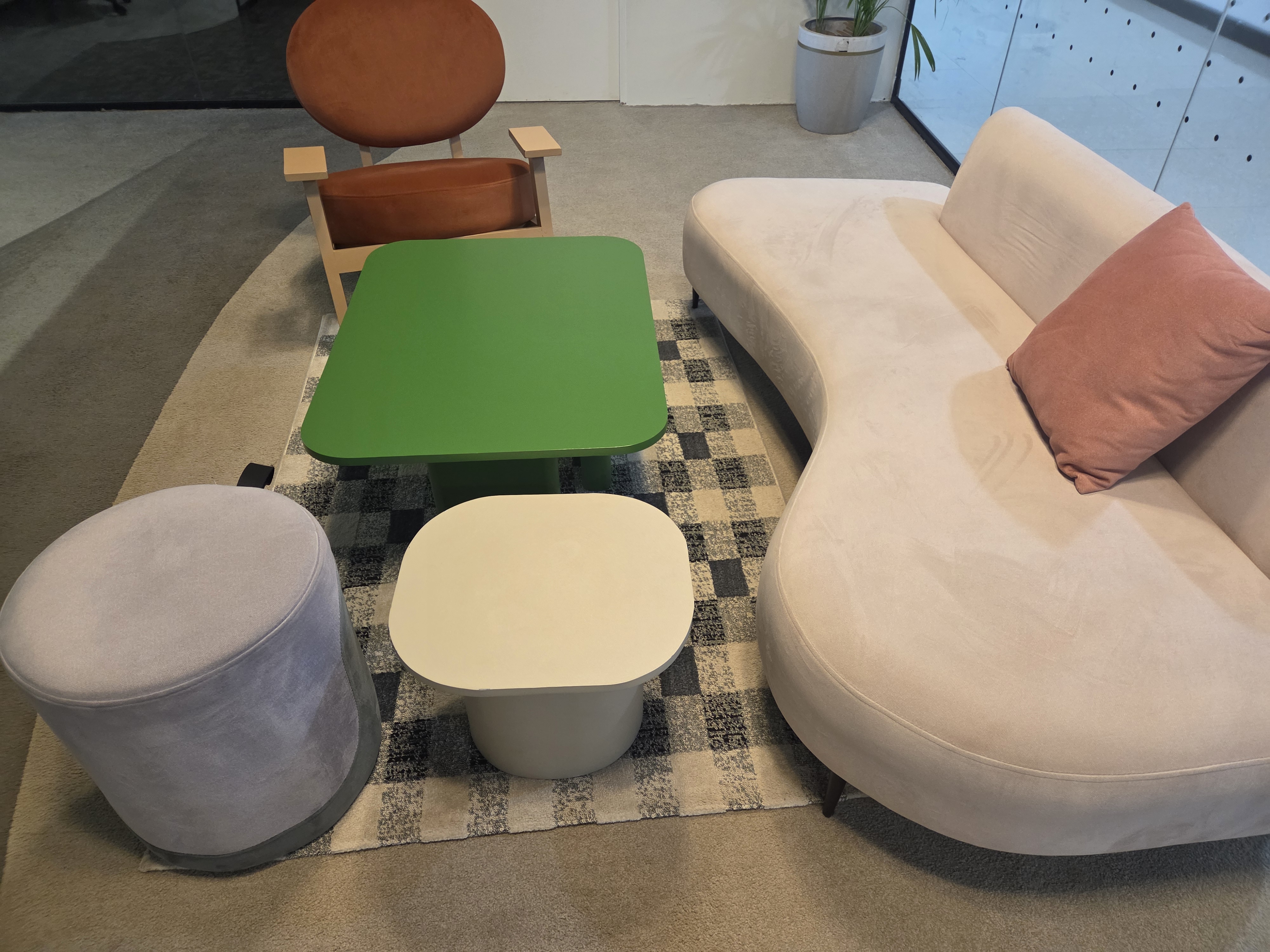 A cozy seating area featuring a modern, curvy beige sofa with a pink pillow, a vibrant green coffee table, and two distinct stools—one round and upholstered in soft lavender, the other an angular white table.