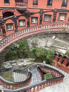 A view of a winding staircase that descends into a rocky, green area. The staircase features ornate railings and balustrades, with intricately carved figures adorning the walls beside it