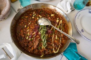 A large, shallow metal pan filled with a rich, dark dish featuring chunks of meat, rice, and vegetables. The dish is garnished with bright green chives, slices of red onion, and a sprig of rosemary, while a shiny spoon rests on the edge.