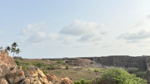 A panoramic view of a rocky landscape with scattered greenery, featuring remains of stone walls and structures in the distance. 
