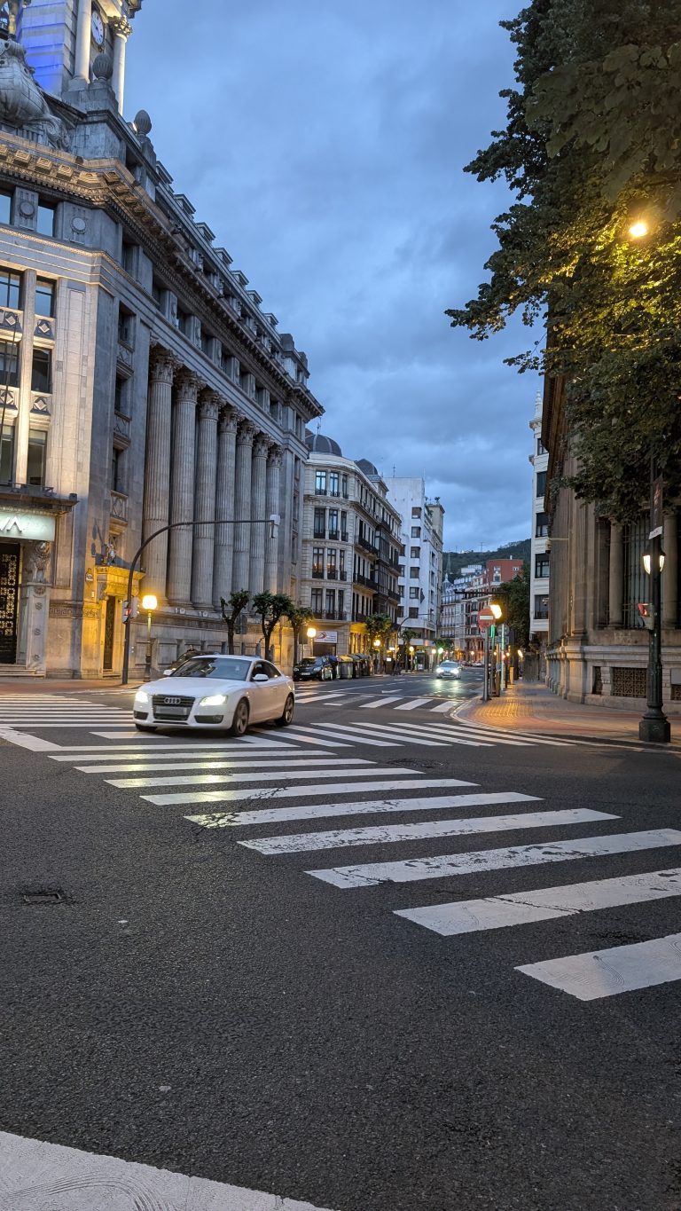 A street scene at dusk featuring a white car driving along a road with zebra crossings. The architecture includes grand buildings with columns, and it appears to be an urban area.