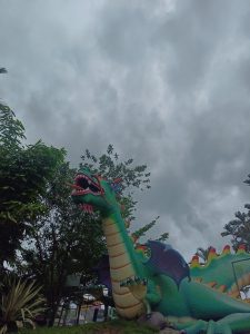 A large, colorful dragon sculpture with a green body, red spines, and black wings is positioned among lush greenery under a cloudy sky. 