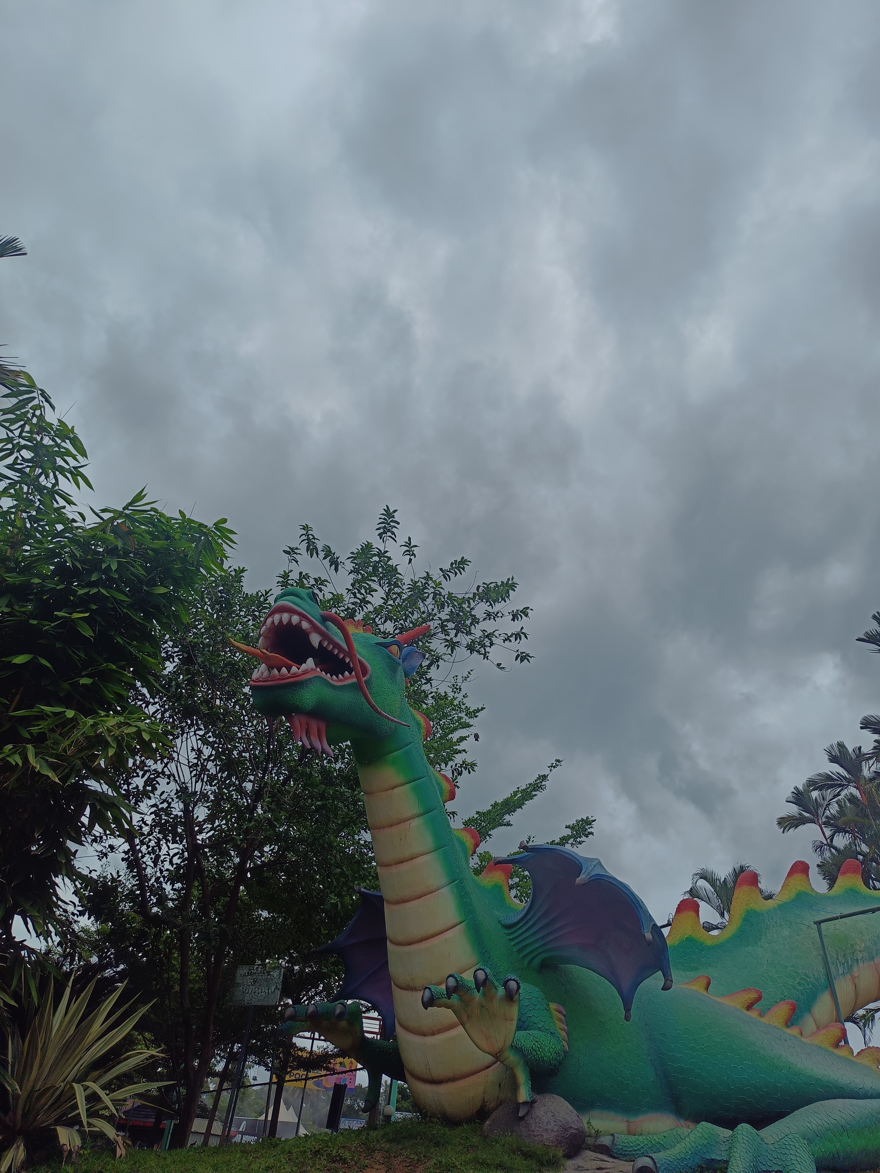 A large, colorful dragon sculpture with a green body, red spines, and black wings is positioned among lush greenery under a cloudy sky.