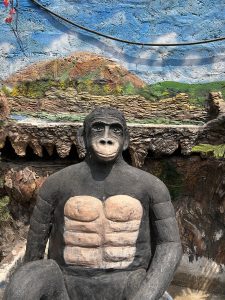 A life-sized sculpture of a muscular chimpanzee with a neutral expression is seated in front of a colorful mural depicting a mountainous landscape and blue sky.