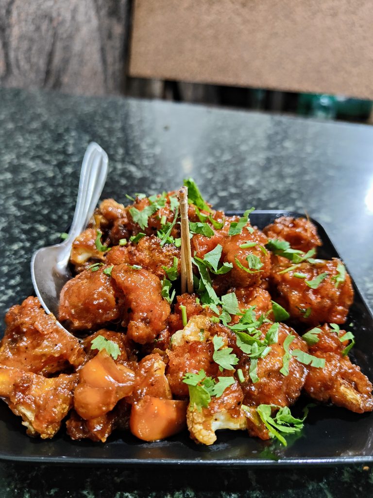 Close-up of Gobi Manchurian — crispy battered cauliflower in a glossy reddish-orange sauce, topped with fresh green cilantro.