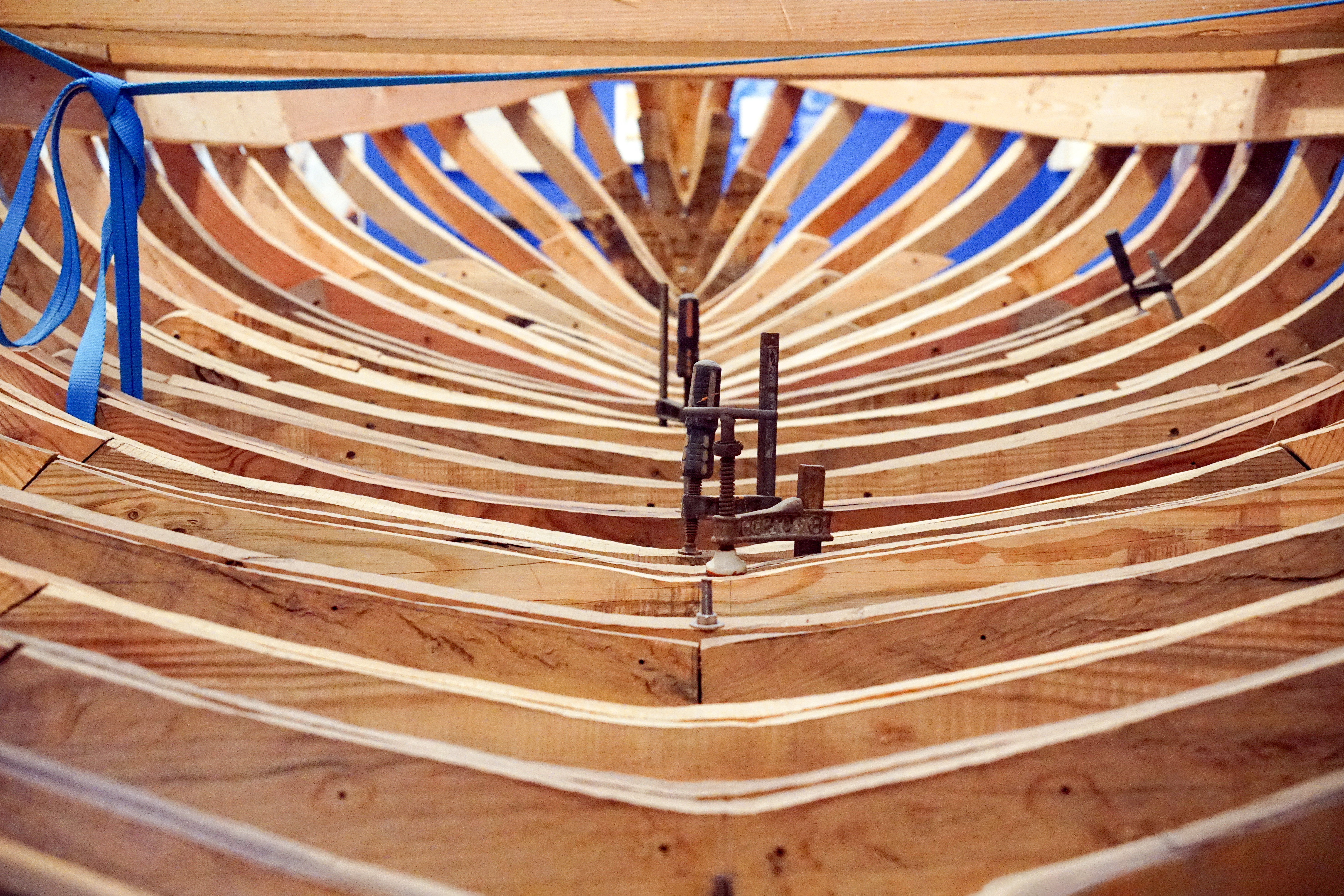 A close-up view of wooden boat frames being assembled, showcasing the curved wooden strips arranged horizontally and vertically within the structure. Metal clamps are securing the pieces together, and blue straps provide additional support.