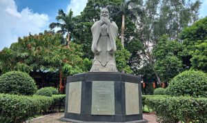 A large stone statue of a contemplative figure stands on a pedestal in a lush green garden.