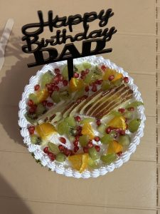 A round birthday cake topped with whipped cream, colorful fruits, and a black cake topper reading “Happy Birthday DAD”, with piped edges on a plain surface.
