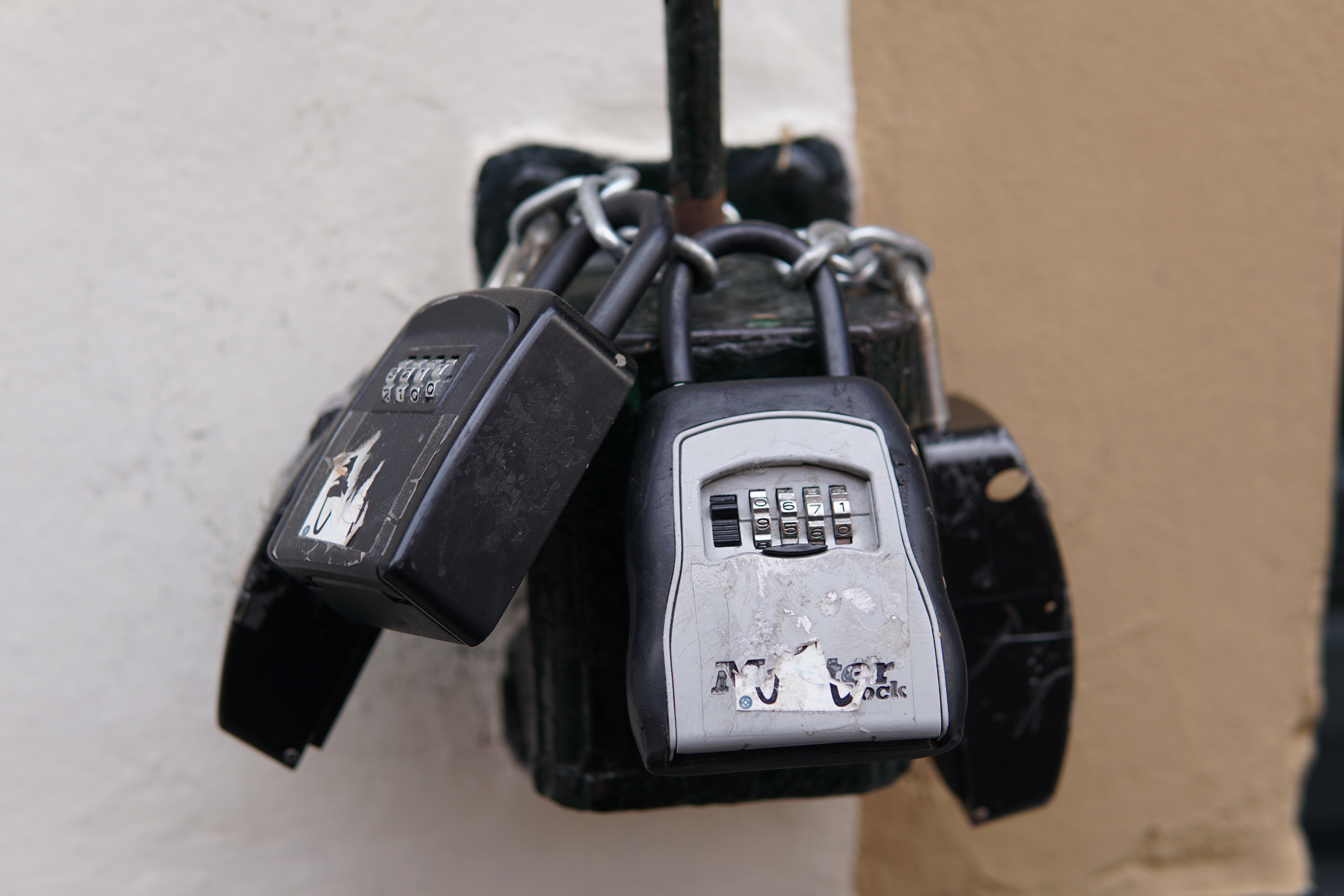 A close-up image of three combination locks attached to a locked metal box with a chain. The locks are a mix of black and gray, showing signs of wear and tear, including scratches and peeling stickers.