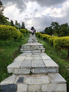 "Two people walk up a stone staircase surrounded by lush green grass and neatly trimmed bushes.