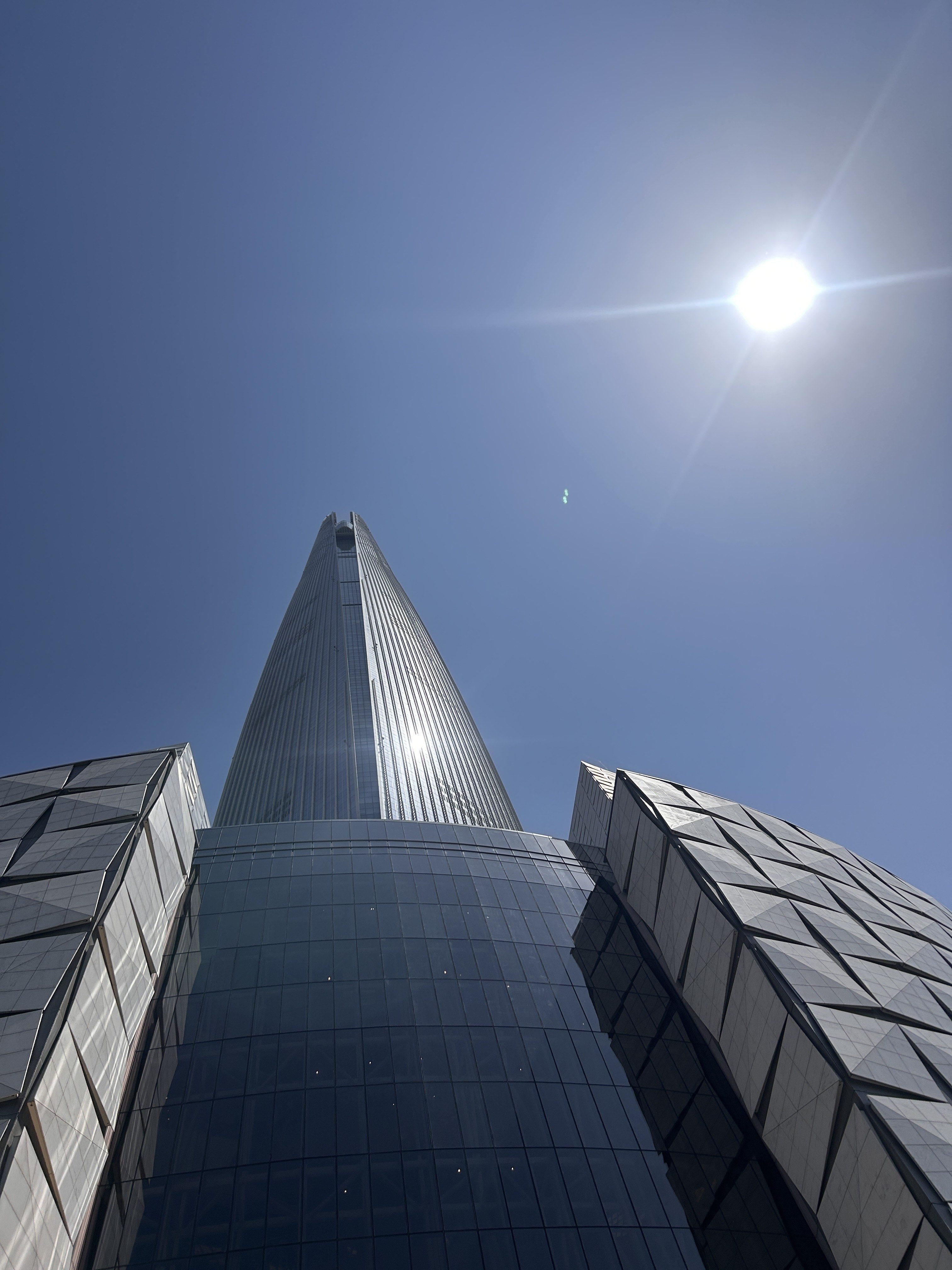 A tall, modern skyscraper towers upward against a clear blue sky, reflecting sunlight on its glass facade. The building's design features angular shapes and competing textures