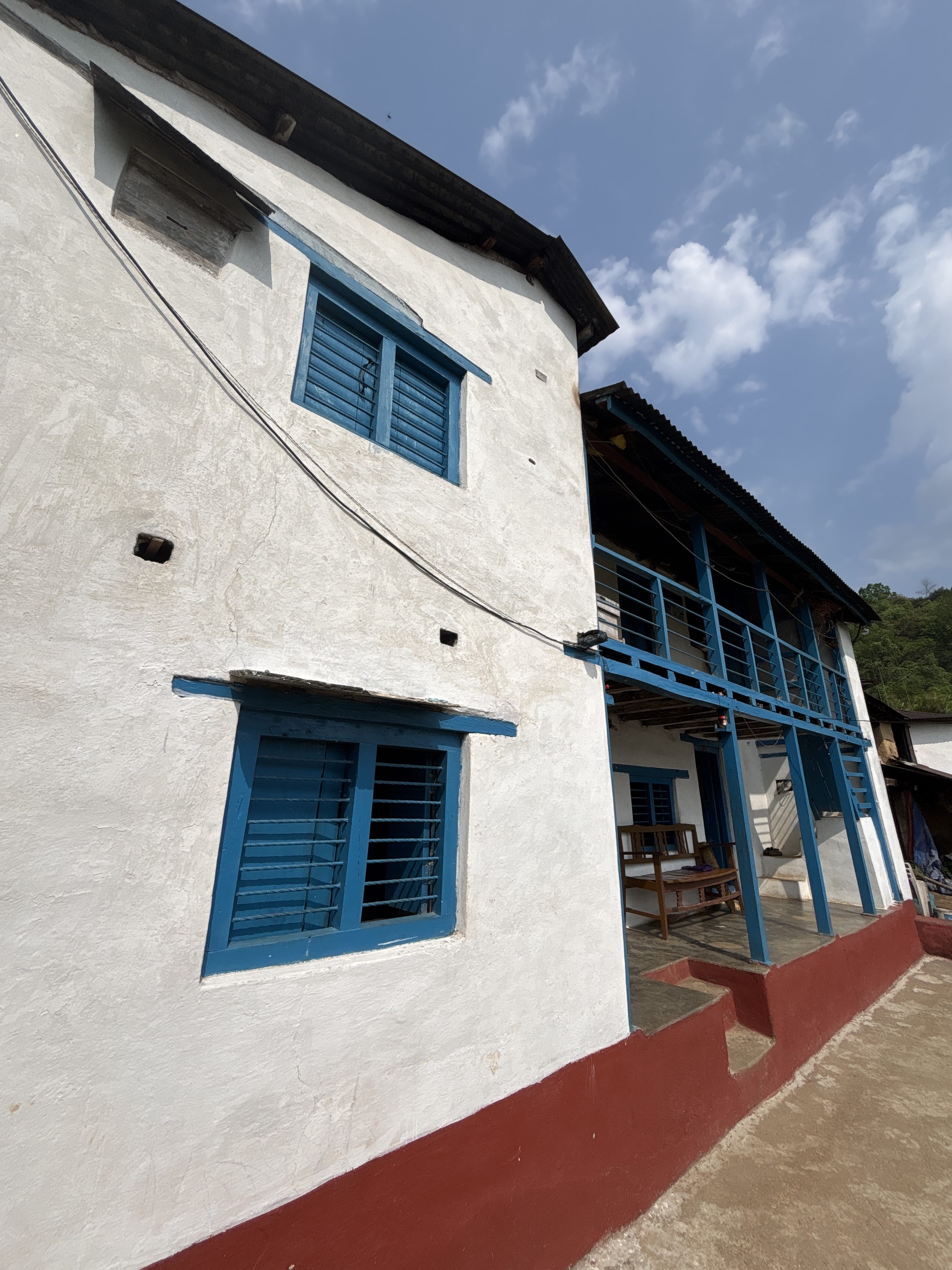 A two-story building with white walls and blue window frames. The exterior features multiple blue-shuttered windows, and the lower section has a red painted border. A staircase leads up to a covered porch with a wooden table and chairs.