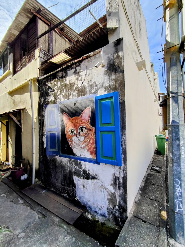 A lifelike mural of a curious orange cat peeking through bright blue painted window shutters on a weathered wall in a narrow alley of George Town, Penang, Malaysia. A charming piece of the city’s famous street art scene.
Note to moderators: Location adds cultural and geographical context. Please keep it.