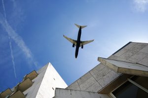 A low-angle view of a commercial airplane flying overhead against a clear blue sky, with wispy clouds visible. The foreground features the angular architectural lines of a modern building made of light-colored stone.