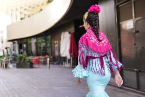 A woman walks away, wearing a traditional outfit that features a light blue dress with ruffles and a vibrant pink shawl adorned with colorful embroidery. Her long hair is braided and decorated with a pink flower.
