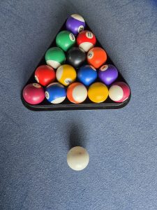 A triangular rack filled with colorful billiard balls sits on a felt pool table. The balls include various colors and numbers, with a solid white cue ball positioned in front of the rack. The background is a blue felt surface.