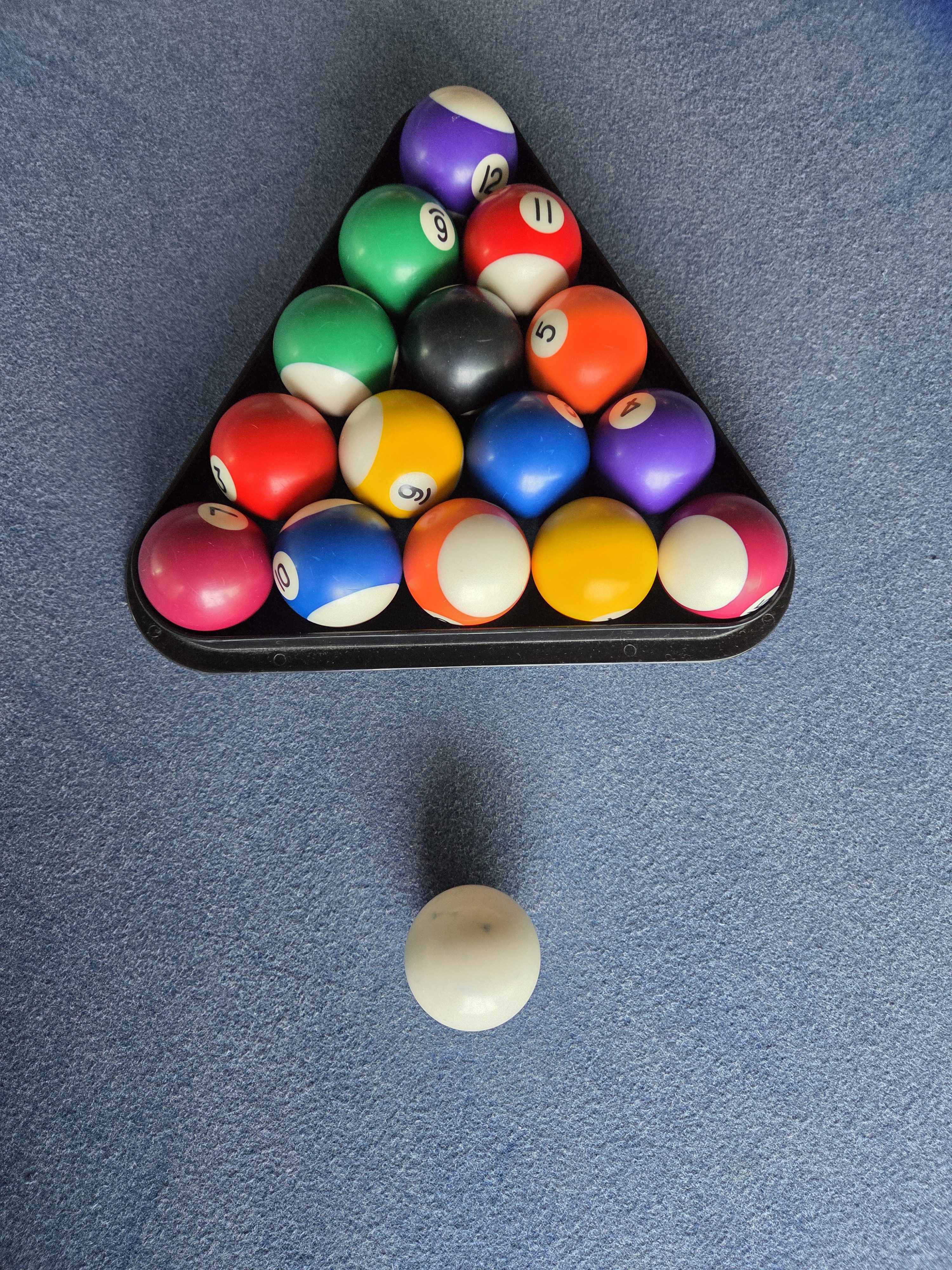 A triangular rack filled with colorful billiard balls sits on a felt pool table. The balls include various colors and numbers, with a solid white cue ball positioned in front of the rack. The background is a blue felt surface.