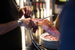 A person is skillfully slicing ibérico ha using a long knife, with their hands visible holding the meat in place. The setting appears to be a food preparation area, with neatly stacked plates in the background.