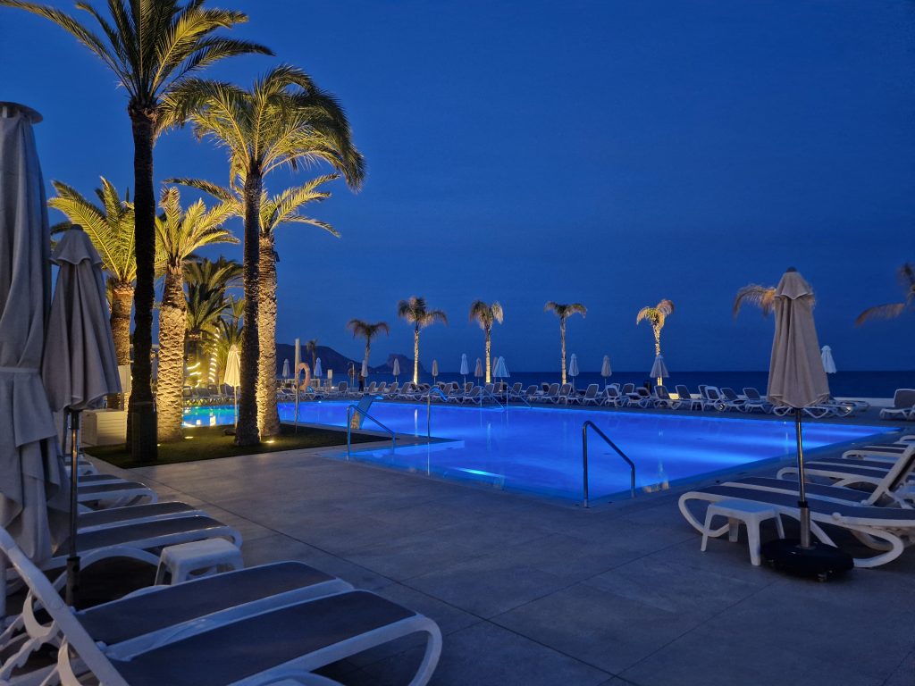 A serene evening view of a seaside resort pool, illuminated in blue and surrounded by palm trees and empty loungers. The calm sea and warm lights create a peaceful atmosphere.