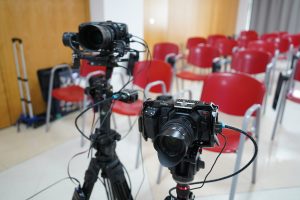 A close-up view of two mounted cameras on tripods, with various cables connected. The cameras are positioned in a room with red chairs in the background.