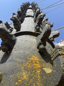The image shows a tall, intricately carved stone pillar with decorative elements along its surface. The pillar is viewed from a low angle, emphasizing its height against a clear blue sky
