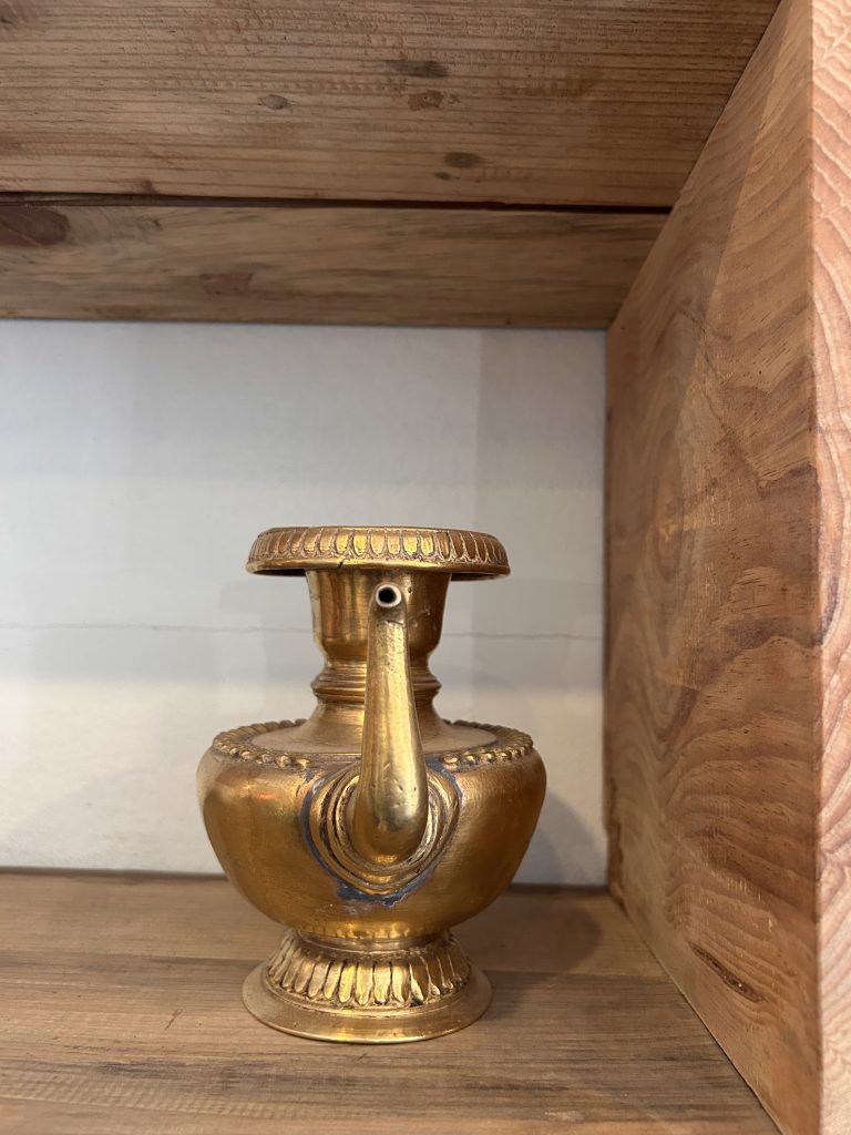 A decorative golden pot with a long spout and ornate detailing, positioned on a wooden shelf.