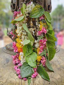 A statue of Lord Shiva, adorned with a garland of colorful flowers and green leaves. The statue is made of a golden-colored metal and is placed against a blurred background. Captured from Vadakkunnathan Temple, Thrissur, Kerala.