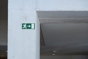 A green emergency exit sign is mounted on a concrete wall, depicting a white silhouette of a person running towards an open door with an arrow pointing to the right.
