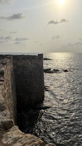 A stone structure extends into the sea, with the sun casting a reflective glow on the water. 
