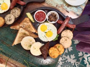 A wooden platter displaying a hearty breakfast featuring two fried eggs with runny yolks on a bed of lettuce, alongside sliced sausages, toast triangles, and various spiced cakes and biscuits. Small bowls hold a red sauce and sautéed mushrooms.